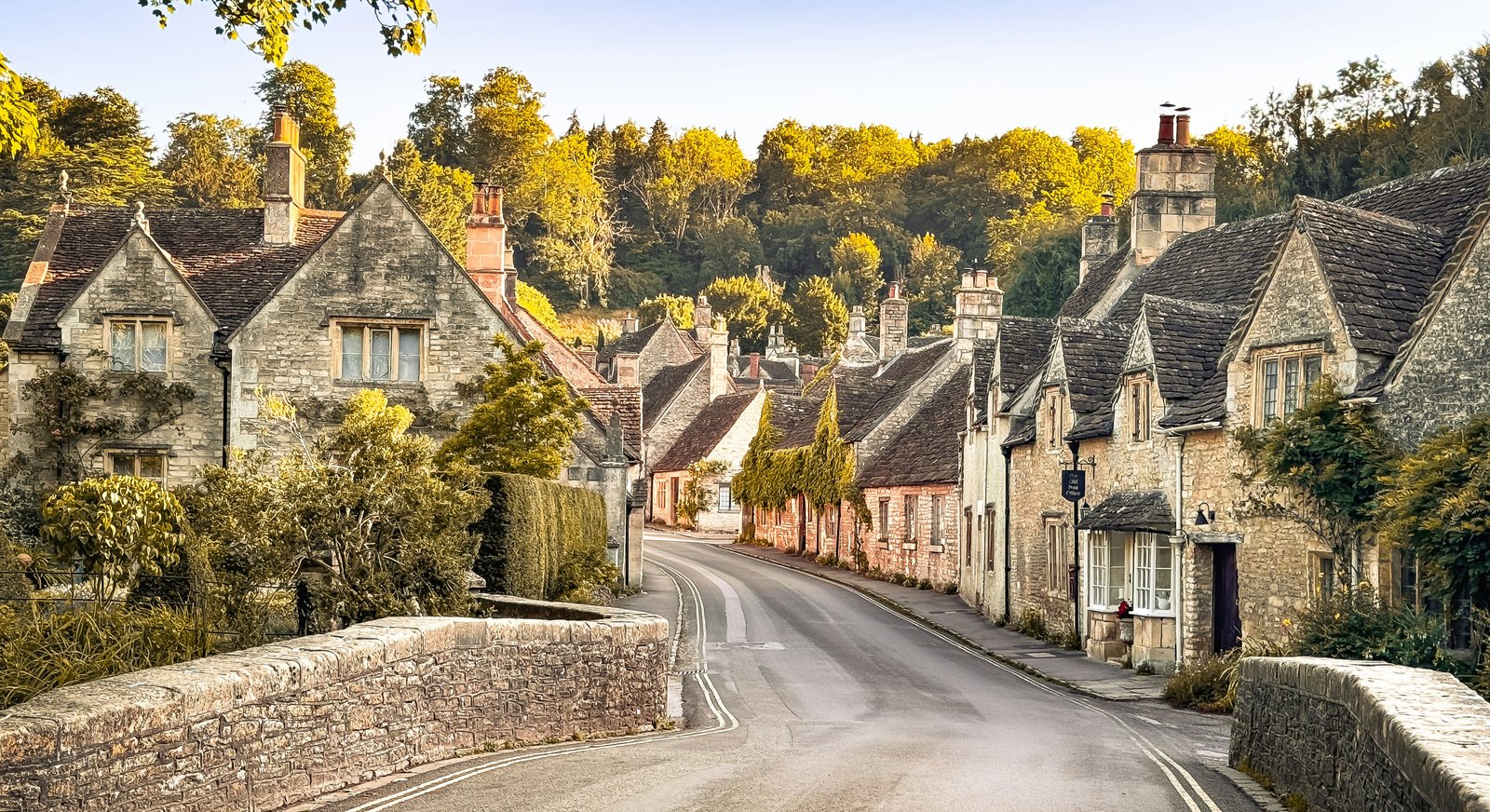 A Cotswolds village high street at golden hour, with honey-colored limestone cottages and a stone bridge.